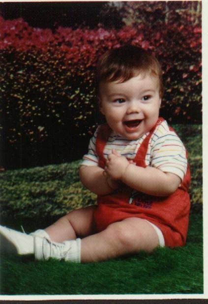 Scott as a toddler, sitting in a garden in red overalls.